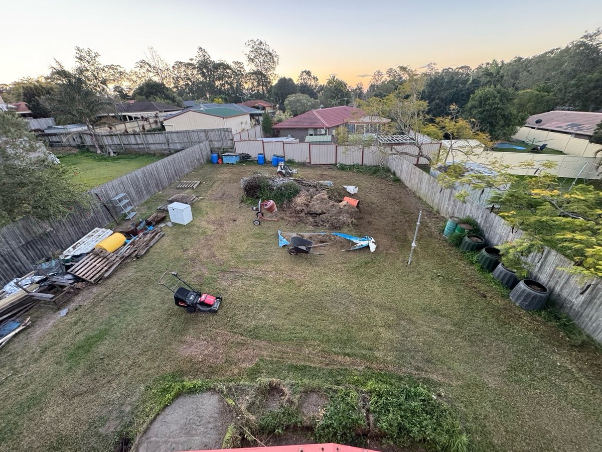 16 Aug 2026 at dusk, seen from a roof: a mown backyard with a lawn mower, wheelbarrow, pallets and a pile of green waste.