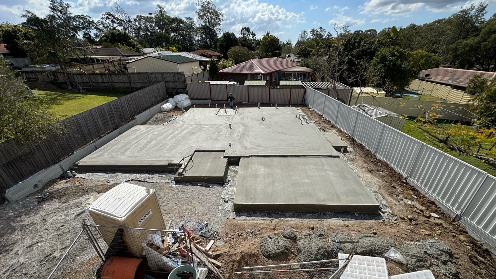 A new concrete slab in a fenced backyard, seen from a roof, with a portable toilet, site fencing and white pods in the foreground